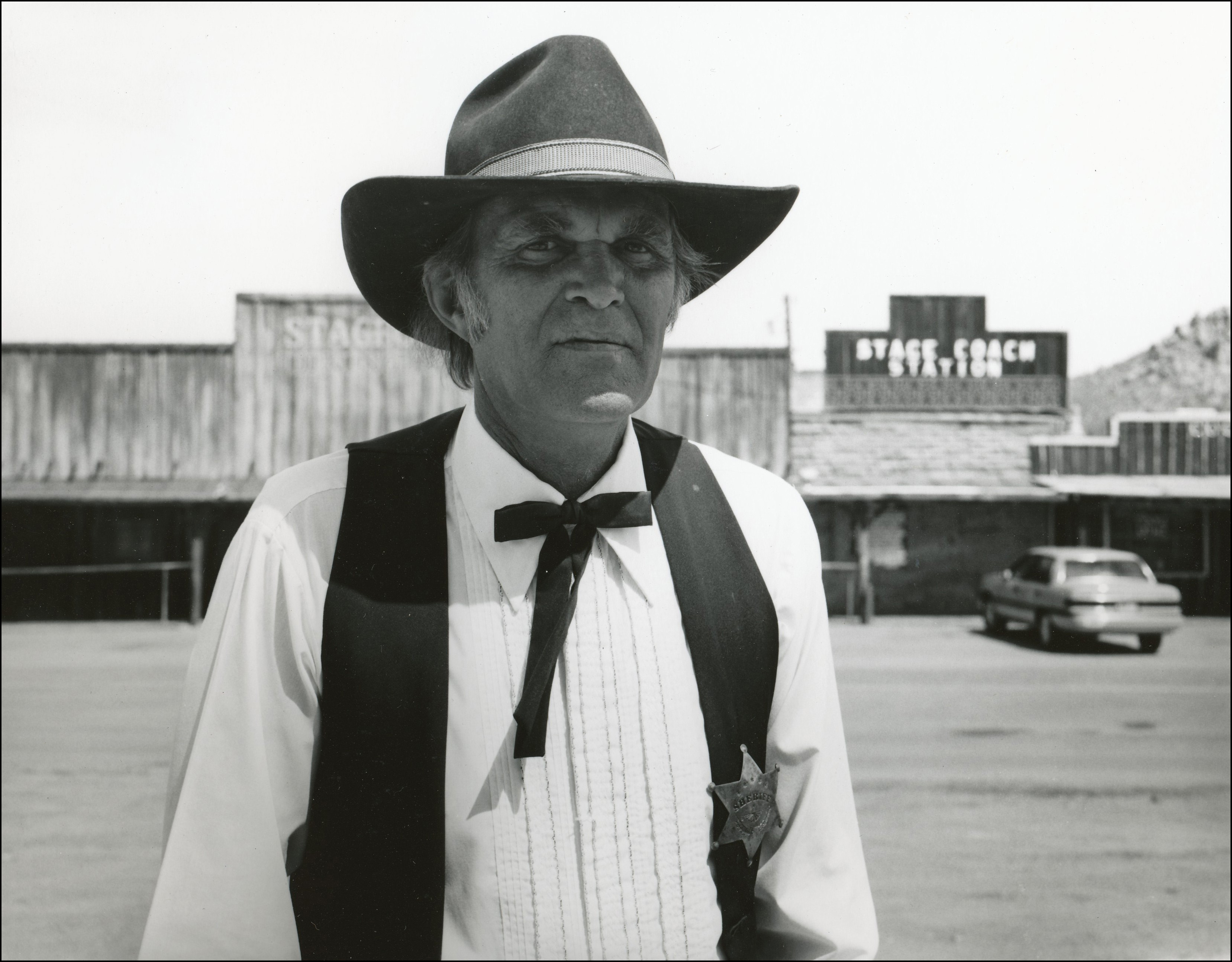 Man with old west clothes on and a sherrif badge with old west buildings in the background